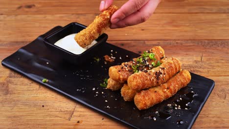 close up of a plate of cheese fingers breaded with sesame seeds, habanero peppers, ranch dressing and worcestershire sauce, a hand takes a cheese finger and dips it in the dressing