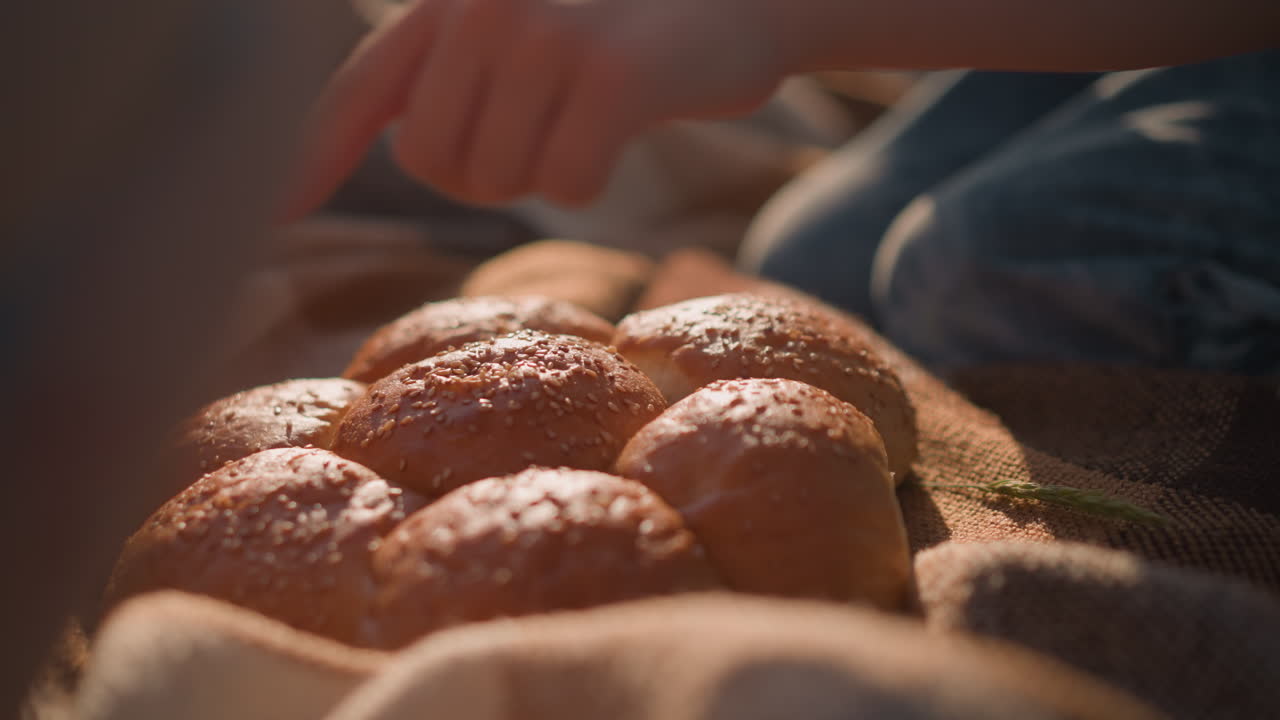 A close-up of freshly baked sesame seed bread rolls placed on a blanket in a grassy field. Children's hands are seen reaching towards the bread, creating a warm and inviting picnic atmosphere