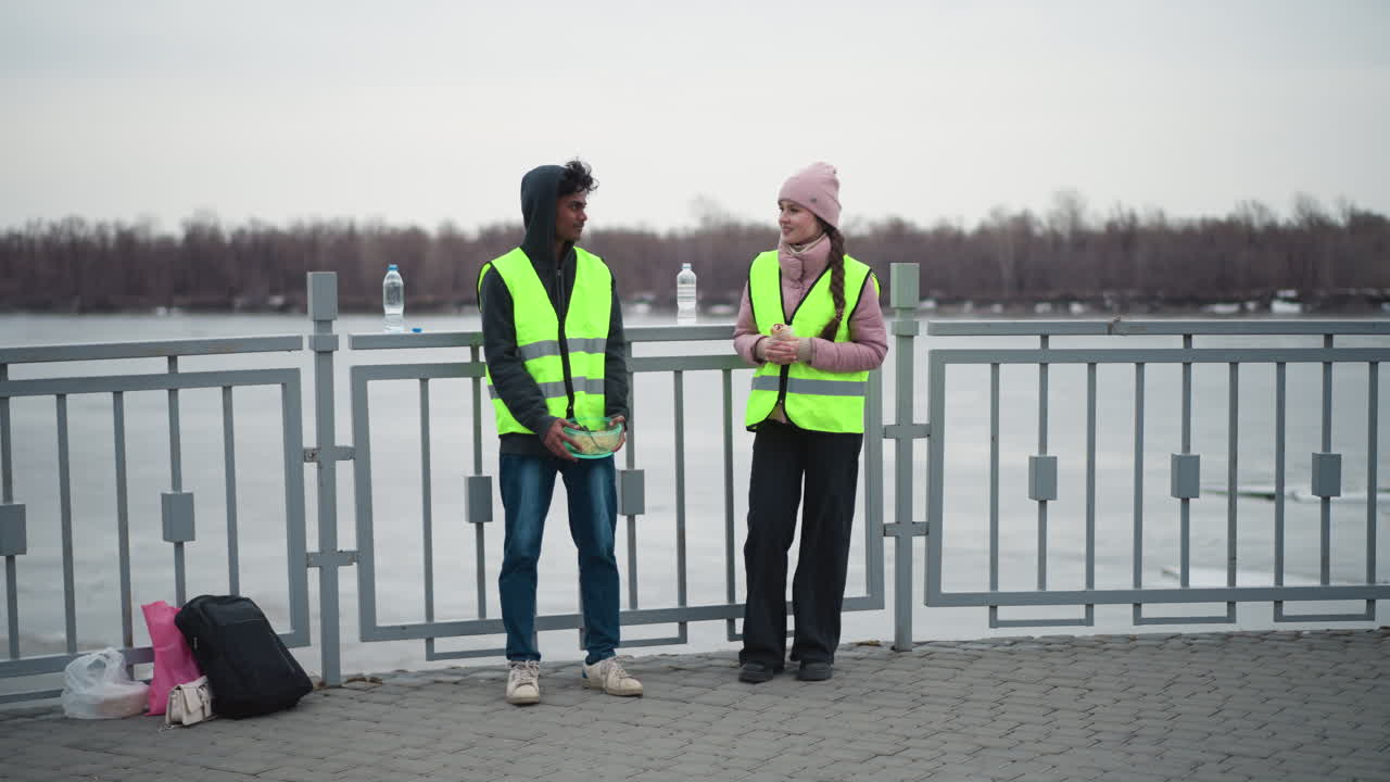 Man in hoodie and neon safety vest holding container of food standing next to woman in pink jacket and matching vest holding wrap, talking on riverside promenade near railing during cold overcast day