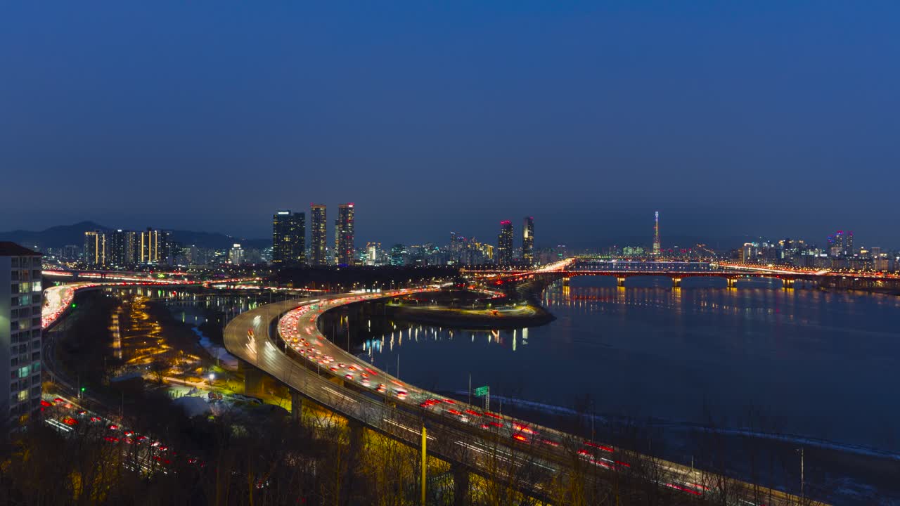 Visuale dall'alto del traffico intenso sulla tortuosa superstrada Gangbyeonbuk-ro con il ponte Seongsu e la Lotte World Tower mentre l'ora blu si trasforma in cielo notturno nel paesaggio urbano di Seul