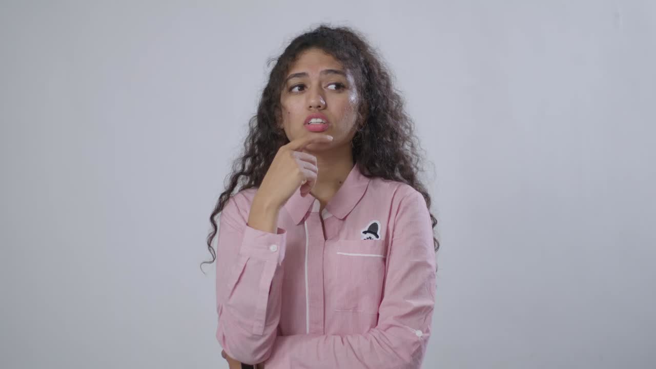 A South Asian young woman in a pink striped shirt rests her chin on her hand, looking thoughtfully against gray background