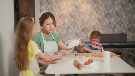 little girl supports sieve for mum pouring flour into bowl while boy at side wears pink toy rings on neck and wrist with baking tools on table in kitchen filled with cozy family activity