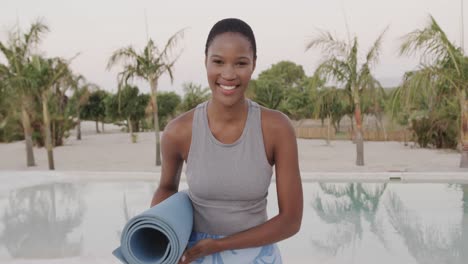 Portrait-of-smiling-biracial-woman-holding-rolled-up-mat-after-practicing-yoga-on-beach,-slow-motion