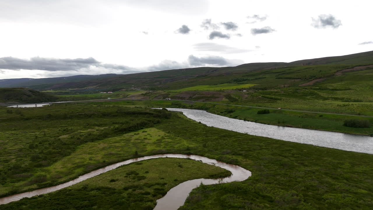 Aerial view of the Skjálfandafljót River winding through wide green valleys toward Skjálfandi Bay, surrounded by scattered farms, rugged mountains and shifting clouds in northern Iceland
