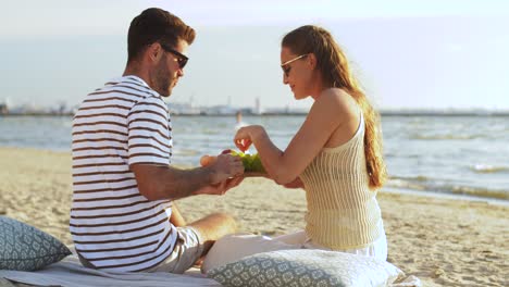 Pareja-Feliz-Con-Comida-Haciendo-Un-Picnic-En-La-Playa.-Concepto-De-Ocio,-Relaciones-Y-Personas.-Pareja-Feliz-Con-Comida-Comiendo-Uvas-Y-Haciendo-Un-Picnic-En-La-Playa.