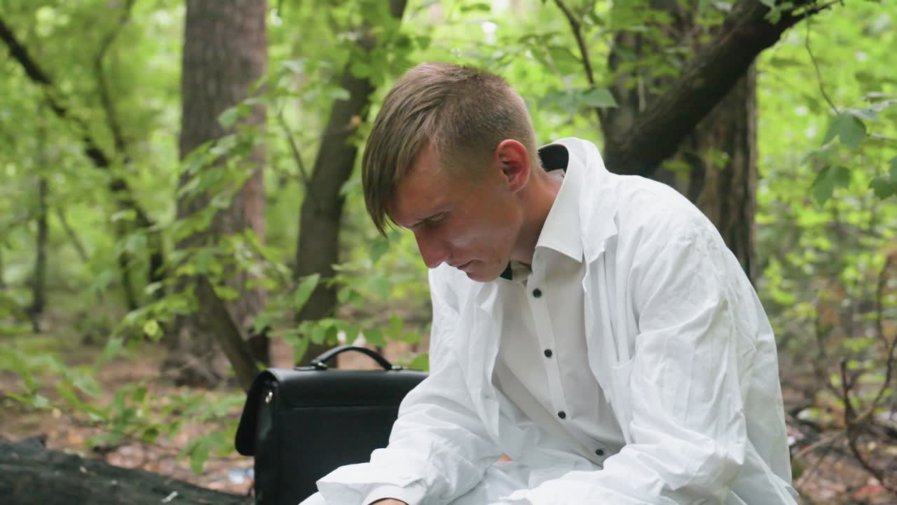 Scientific researcher in white coat sitting on dry stump in forest removing glasses while showing signs of fatigue and exhaustion during outdoor work with bag placed beside him surrounded by trees