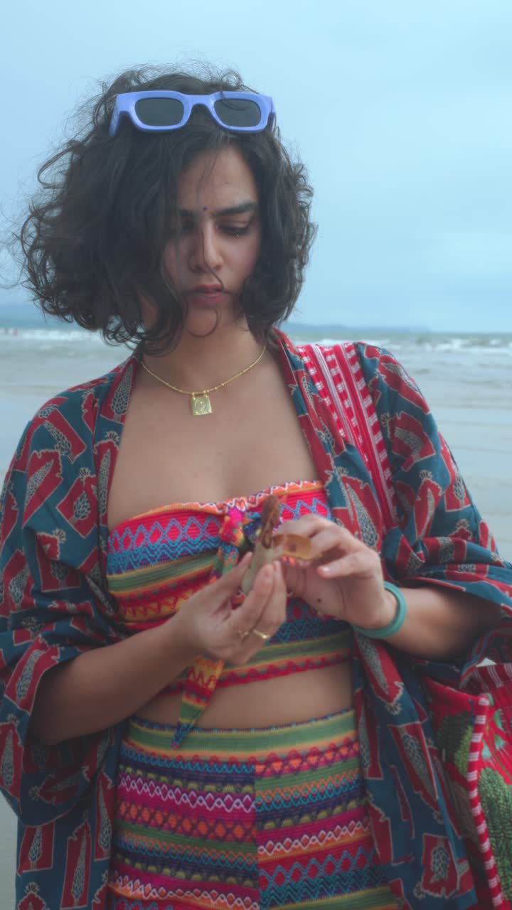Vertical portrait of young woman in vibrant patterned beachwear with sunglasses, necklace and gold pendant, standing on a beach under daylight. Vibrant colors and summer mood
