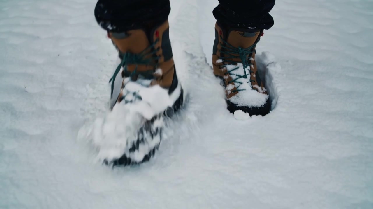 Excursionista solitario cambiando el peso y pisando suelo nevado, dejando huellas de botas color canela con cordones verdes visibles.