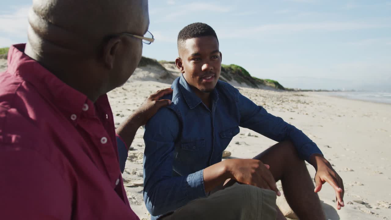 Ayah Afrika-Amerika duduk di pantai bersama putra remajanya, meletakkan tangan di pundaknya, dan berbicara.