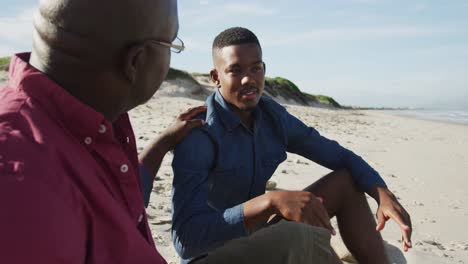 padre afroamericano sentado en la playa con su hijo adolescente, poniendo la mano en su hombro y hablando