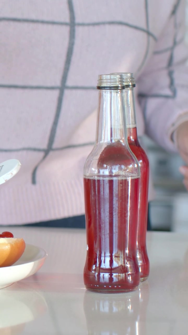 Video verticale: Uomo che si allunga con maglietta a griglia rosa, spingendo una bottiglia di salsa in cucina per la preparazione vicino a un mezzo panino