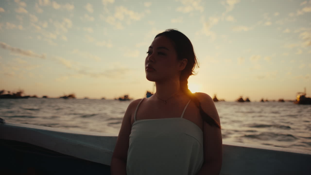 Young woman sitting on boat deck looking toward horizon over calm sea during warm evening light, relaxing calm travel