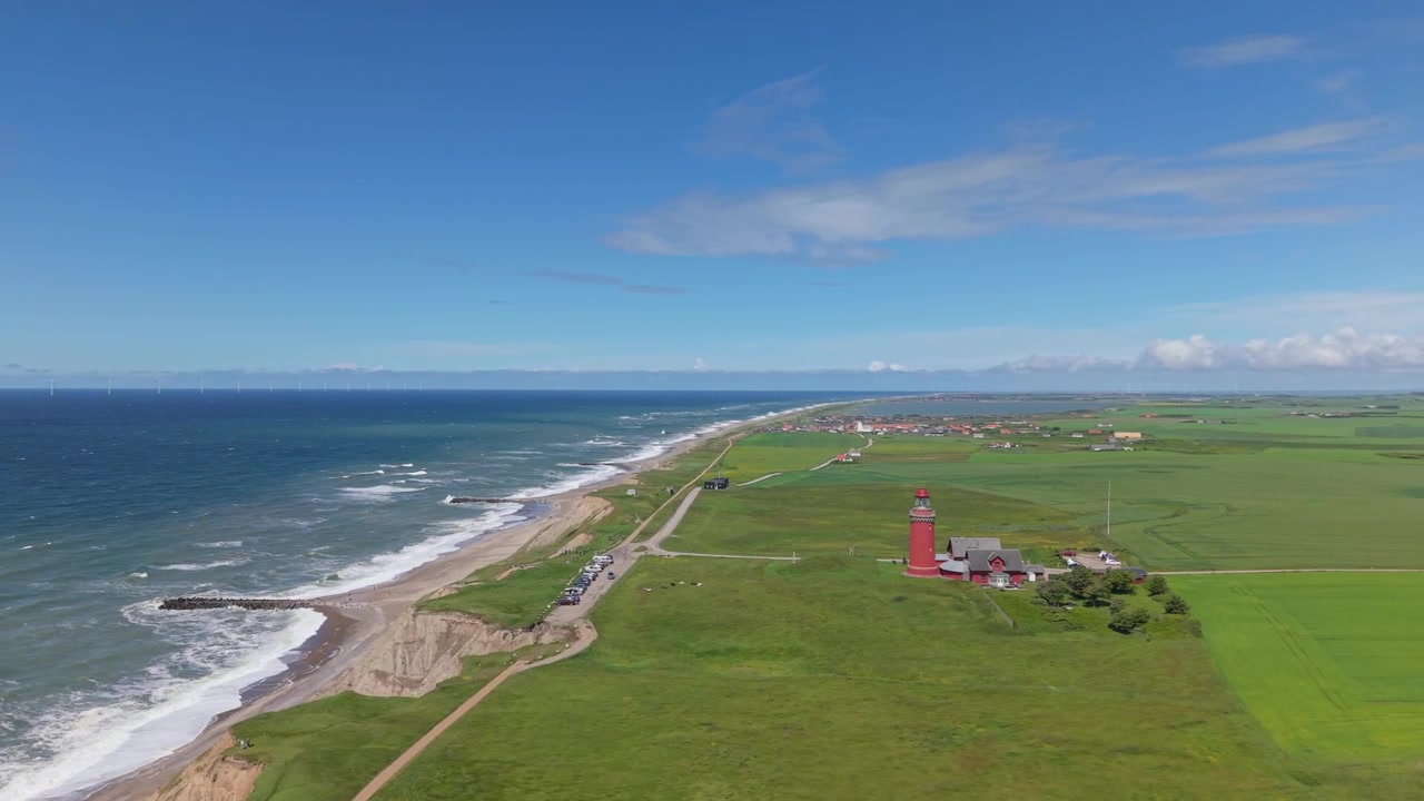Panoramisk flygbild som visar Västra Jyllands natursköna kustlinje, med den ikoniska röda Bovbjerg fyr omgiven av frodigt grönt gräs och Nordsjöns blå vatten