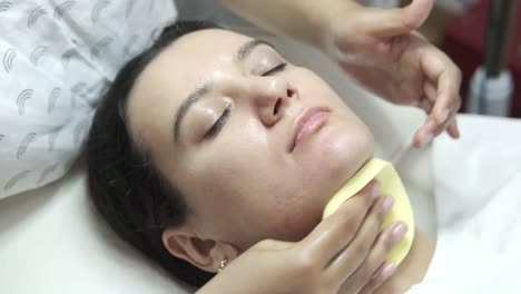 relaxed woman lying in salon and having deep cleansing nourishing facial treatment. beautician cleaning client face with round sponge. healthcare, wellness and medicine