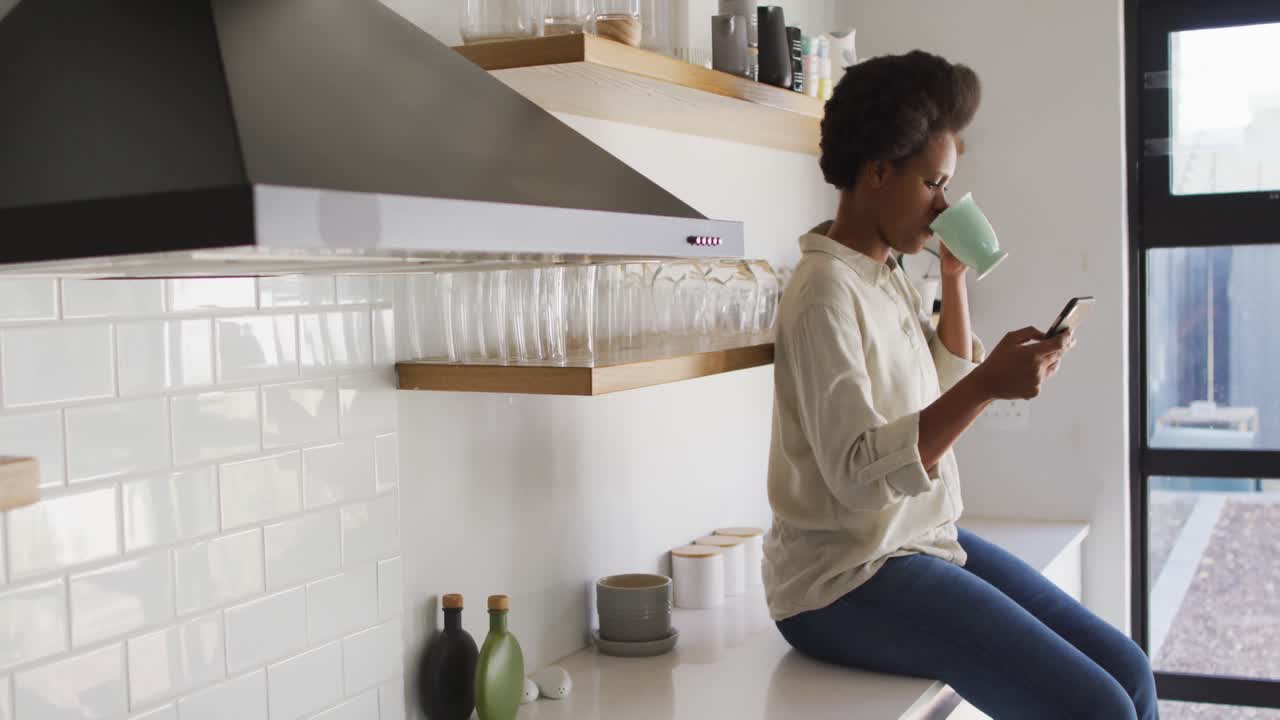 feliz mujer afroamericana bebiendo café y usando un teléfono inteligente en la cocina