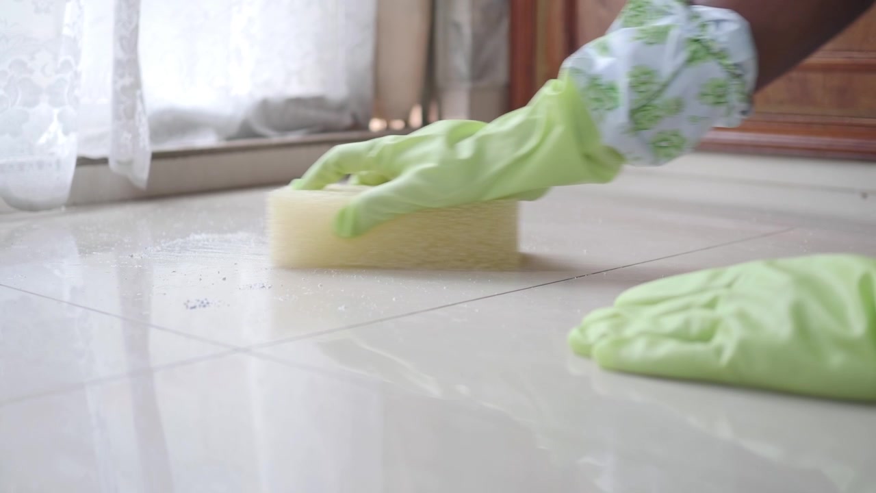 Persona che indossa guanti di gomma e pulisce un pavimento piastrellato con una spugna