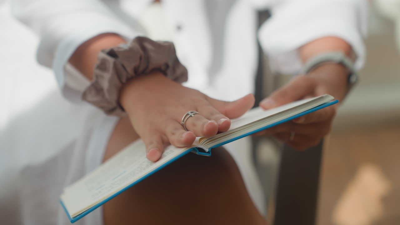 Mujer leyendo un diario en una terraza de isla, cuaderno azul sobre rodillas iluminadas por el sol, primer plano de manos con anillos y coletero, pasando páginas lentamente, ambiente relajado de resort, atmósfera íntima de vida lenta
