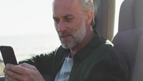 happy caucasian man sitting in car using smartphone on sunny day by the sea