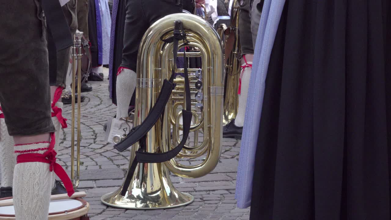 A tuba rests on a cobblestone surface, surrounded by musicians in traditional dress, shown only from the waist down. A quiet moment before the music begins.