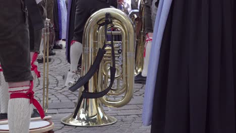 a tuba rests on a cobblestone surface, surrounded by musicians in traditional dress, shown only from the waist down. a quiet moment before the music begins.