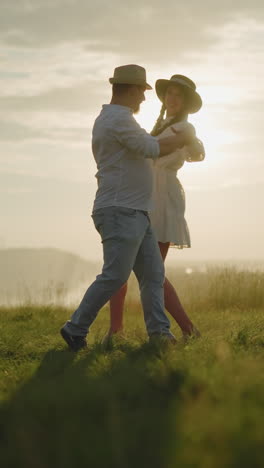 una pareja amorosa baila en una colina cubierta de hierba iluminada por el sol, disfrutando de un momento tierno juntos. el hombre, vestido con una camisa blanca, sombrero y vaqueros, abraza a su esposa, que lleva un vestido blanco fluido