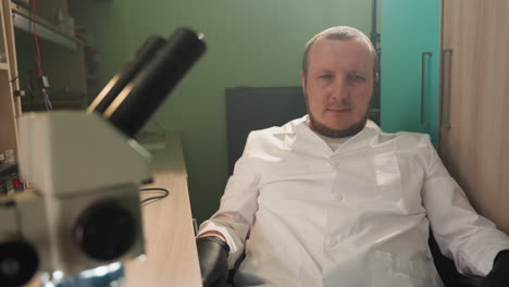 a close-up view of a technician in a white lab coat and black gloves, leaning back and looking content in a lab setting, with a microscope on the table