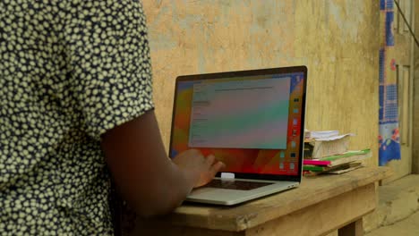 Escribiendo-Con-Una-Computadora,-Una-Estudiante-Está-Ocupada-Con-Sus-Estudios-Justo-En-Frente-De-Su-Casa-En-Un-Pueblo-De-Kumasi,-Ghana,-África