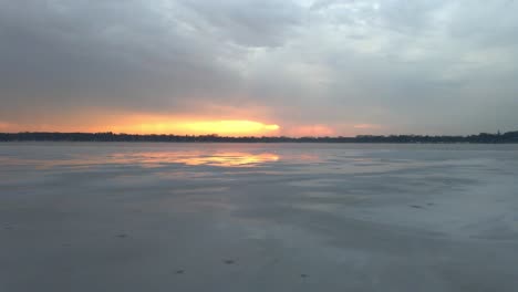 Aerial-view-of-a-frozen-lake-ice-reflecting-the-sky-and-a-colorful-sunset-on-the-horizon,-nature,-beautiful-Minnesota