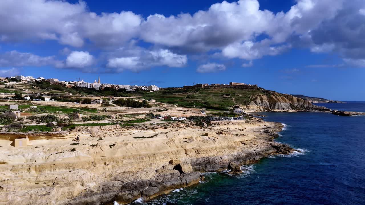 Ampia inquadratura panoramica dall'alto della costa rocciosa calcarea e delle acque turchesi del Mediterraneo di Gozo, Malta, sotto un cielo azzurro brillante con soffici nuvole bianche durante un soleggiato pomeriggio estivo