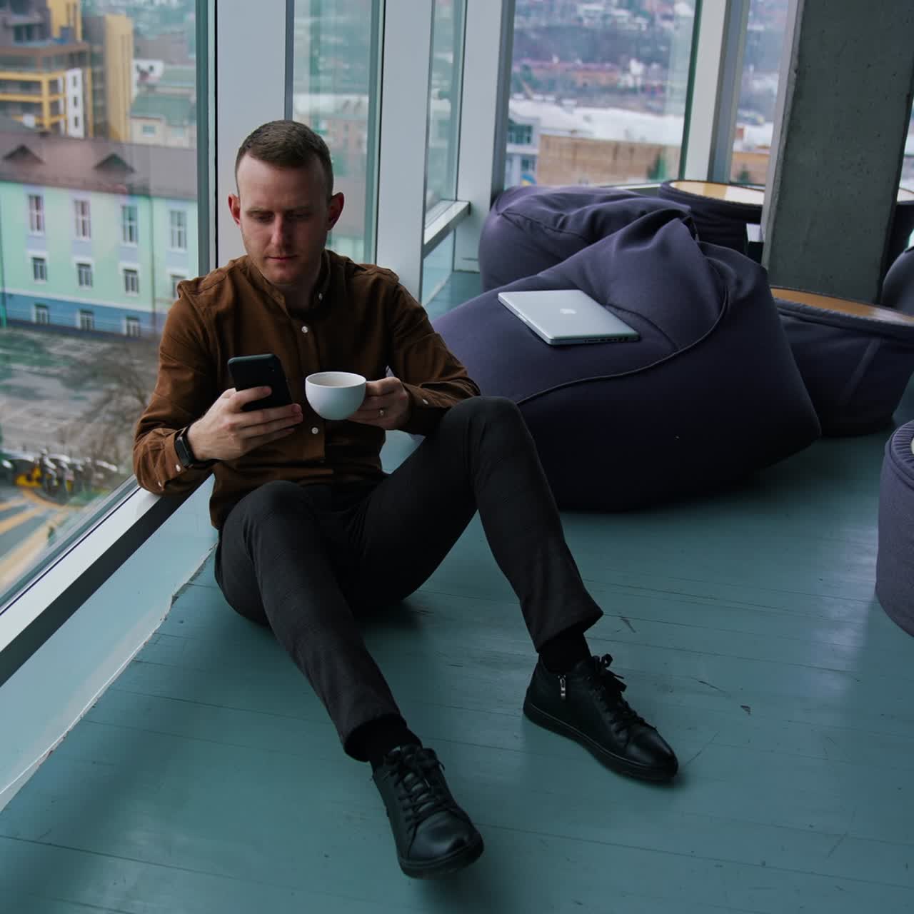 Young businessman having break. Handsome guy in shirt and trousers sitting on a floor with a cup of coffee and a phone near the window in modern office in the city
