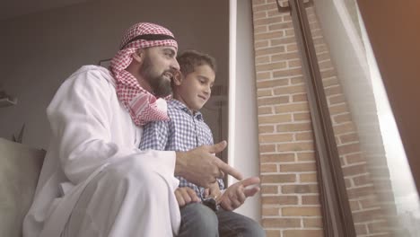 happy father and son standing near the window while father pointing away and smiling. arab family concept.