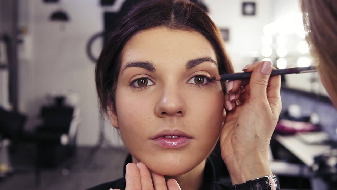 Unrecognizable make up artist's hand applies eyeliner in the eyes' outer V section of a beautiful attractive brunette girl. Beauty salon background.