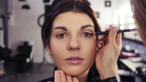 unrecognizable make up artist's hand applies eyeliner in the eyes' outer v section of a beautiful attractive brunette girl. beauty salon background.