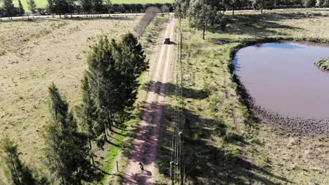 Video-Aereo-De-Un-Auto-Saliendo-De-Una-Casa-En-El-Campo-Mientras-Un-Perro-Lo-Corre,-Con-Un-Lago-Al-Fondo-Y-Muchos-Arboles