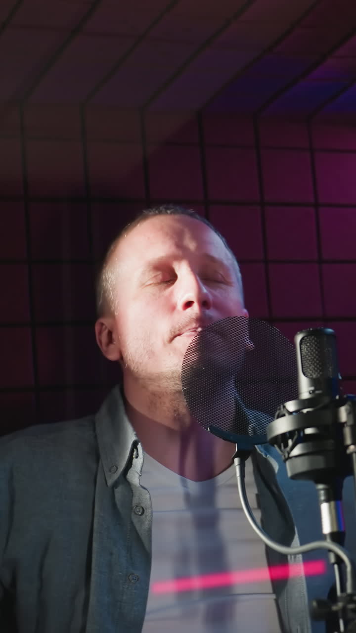 un hombre con una camisa de manga larga azul y camisa interior blanca sostiene un micrófono en un estudio a prueba de sonido rojo, listo para cantar. la iluminación dramática enfatiza la intensa atmósfera de una sesión de grabación