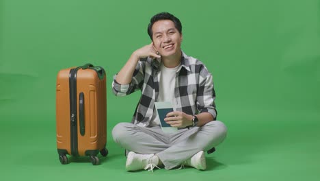 full body of asian male traveler with luggage and passport smiling and making call me gesture while sitting in the green screen background studio