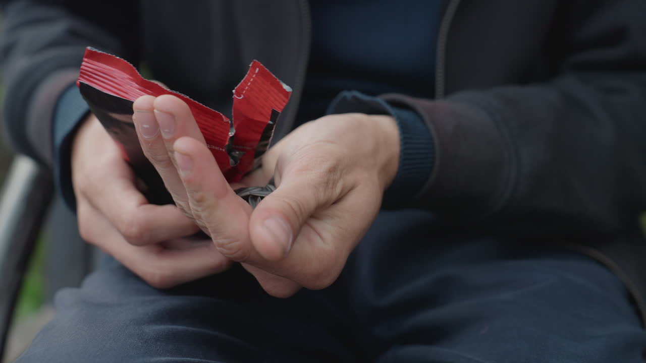hands detail white man unwrapping red wrapper, seated on bench outdoors in casual jacket fingers peeling foil and plastic layers, tactile focus on palms and knuckles, slow careful motions reveal