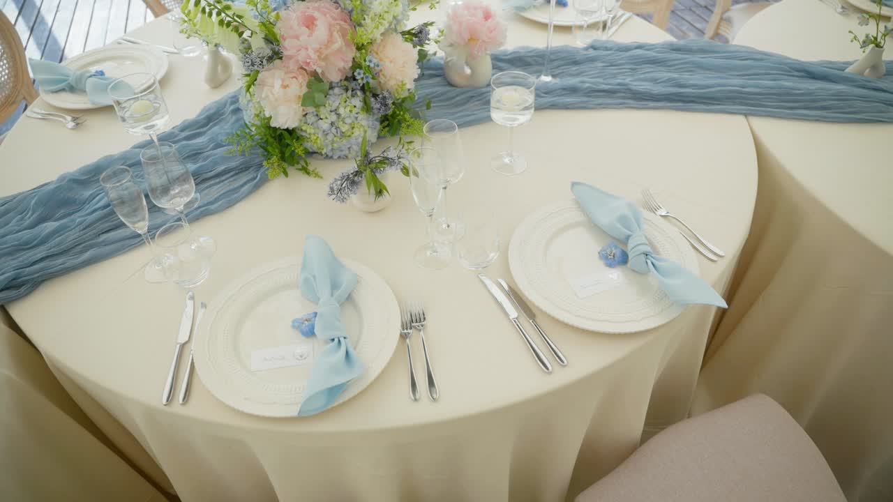 Elegant wedding table set with blue napkins, pastel floral centerpiece, and fine glassware on ivory linen—perfect for a romantic and upscale reception setting