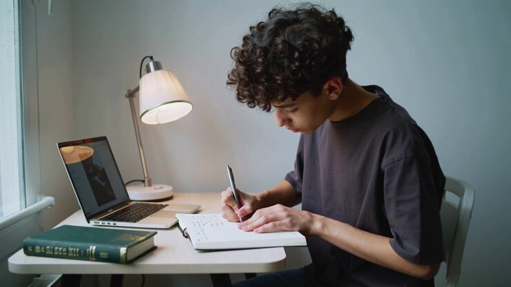 En teenager i mørk T-shirt bladrer i spiralindbundne sider, studerer og skriver ved et skrivebord ved vinduet med en kuglepen og bærbar computer.