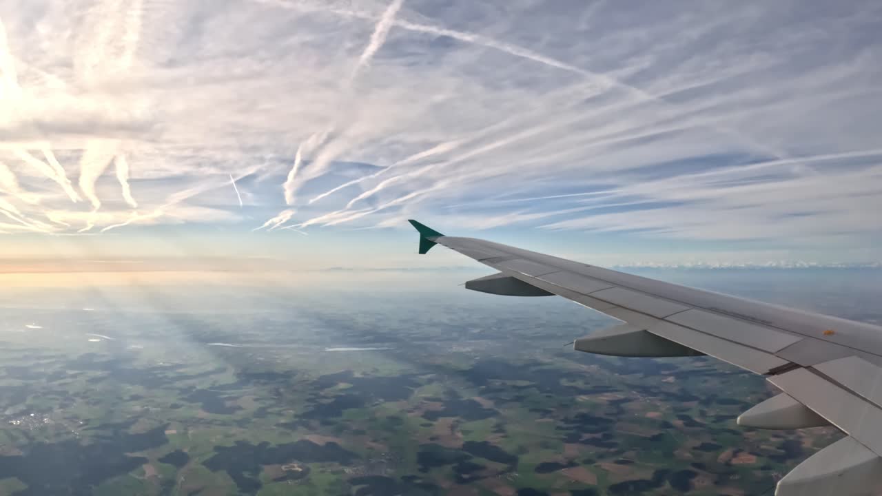View from airplane window showing wing flying over Germany during sunrise, with contrails crossing the blue sky and fields and forests below, creating a scenic travel image