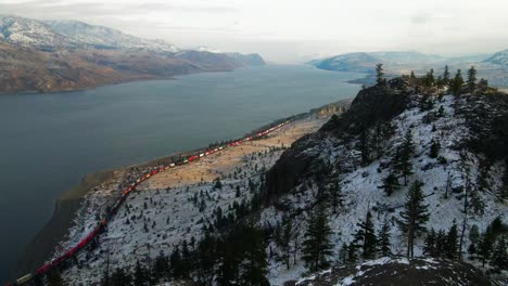 cinematic shot of a colorful train passing by kamloops lake in the winter, snow-covered rocky cliffs and spruce trees are visible in the foreground