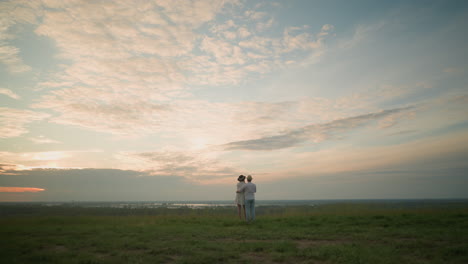 newlyweds stand on a grassy hill beside a tranquil lake at sunset, gazing at the sky. the man, in a white shirt, hat, and jeans, holds the woman by the waist. she wears a black hat and a white dress