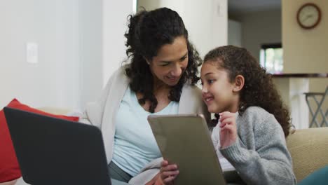 madre e hija de raza mixta felices sentadas en el sofá, divirtiéndose, usando portátil y tableta