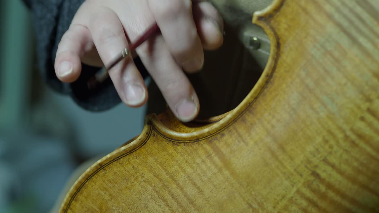 Master violinmaker works on violin ribs using finger tamping and a fine brush, enhancing patina and texture while the marbled back plate, purfling, and worn edges reveal detailed antiquing