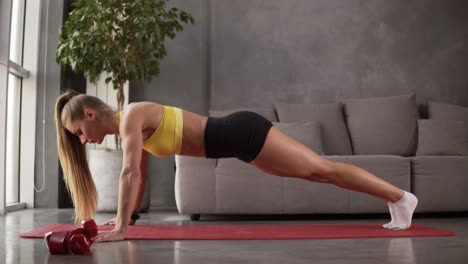 healthy young woman in sportswear - yellow top and black shorts doing push ups on fitness mat in front the grey sofa in living room. young female exercising at home. side view