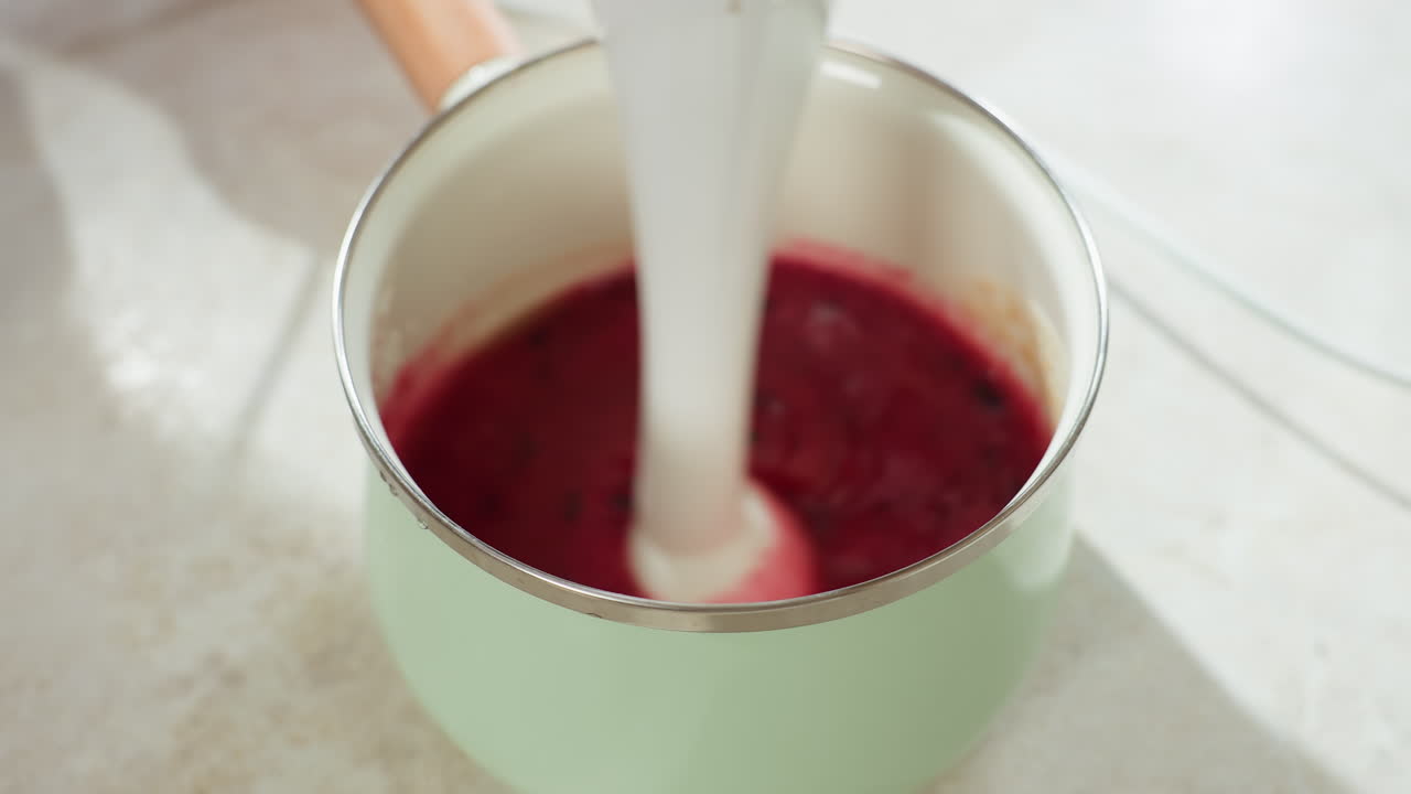Close up of person using electric blender to stir vibrant red currant berry mixture in green pot on kitchen counter during food preparation