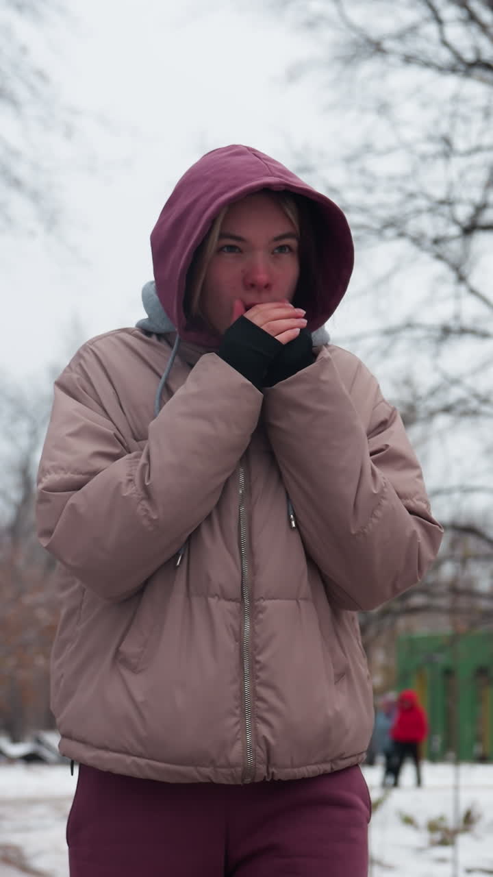 겨울 옷을 입은 평온한 여자가 눈으로 인 공원을 혼자 산책하며, 따뜻함을 얻기 위해 입에 손을 고, 벌거벗은 나무, 어진 눈, 그리고 배경에 흐릿한 인물