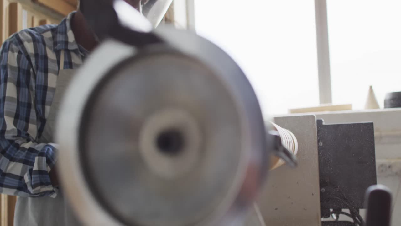 афроамериканский плотник, носящий защитный шлем, превращающий древесину на токарном станке в столярной мастерской