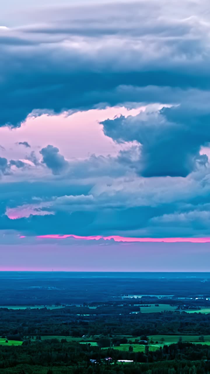 El time-lapse revela unas espectaculares nubes de color azul oscuro con matices rosas que se desplazan sobre el vasto paisaje rural letón, con bosques verdes, campos y un horizonte lejano durante una vibrante puesta de sol.