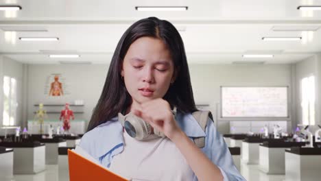 close up of asian teen girl student with a backpack reading book and yawning while standing in science laboratory
